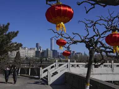 Wanita mengenakan masker untuk membantu melindungi dari virus corona berjalan dengan dekorasi lentera jelang Tahun Baru Imlek di sebuah taman di dekat cakrawala kota di Beijing, Kamis (27/1/2022). (AP Photo/Andy Wong)