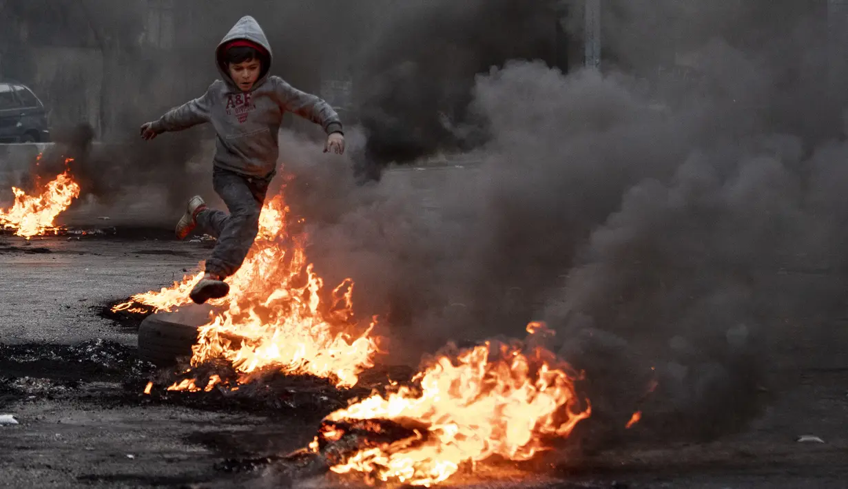Seorang anak laki-laki melompati ban yang dibakar untuk memblokir jalan, selama protes di Beirut, Selasa (2/3/2021). Warga Lebanon yang marah memblokir jalan di seluruh negeri dengan membakar ban dan tempat sampah setelah mata uang Lebanon jatuh ke titik terendah baru. (AP Photo/Hassan Ammar)
