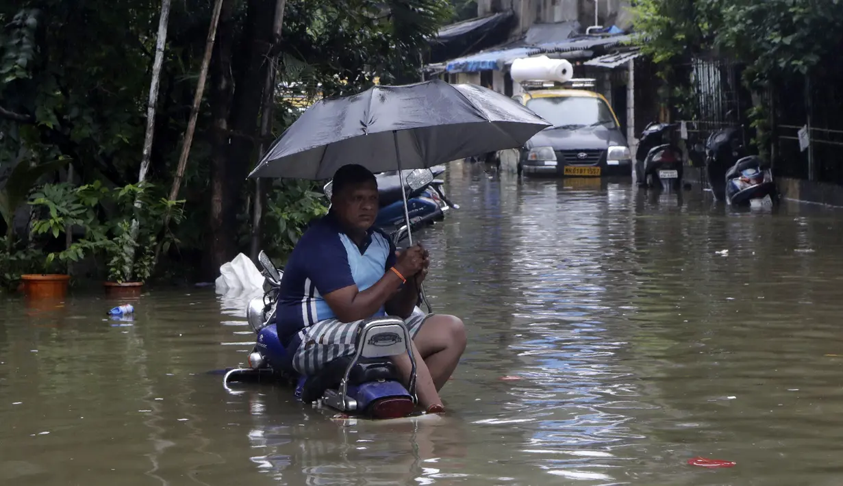 Seorang pria duduk duduk memegang payung di atas sepeda motor yang diparkir di jalan yang tergenang air setelah hujan lebat di Mumbai, India, Rabu (23/9/2020). Musim hujan di India berlangsung dari Juni hingga September. (AP Photo/Rajanish Kakade)