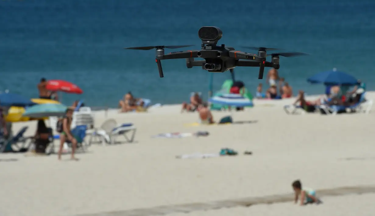Sebuah drone atau pesawat tak berawak yang diterbangkan oleh polisi setempat memperingatkan orang-orang untuk menjaga jarak di pantai Areas di Sanxenxo, Spanyol, Selasa (14/7/2020). Drone tersebut untuk memantau jaga jarak antarwisatawan saat berlibur di masa pandemi Corona. (MIGUEL RIOPA / AFP)