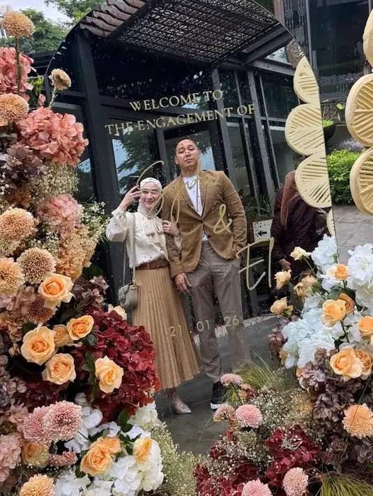 Dalam foto bersama kekasih saat mirror selfie,  Melody tampil feminin dengan blus putih detail pita yang dipadukan rok pleats cokelat muda. Look-nya terlihat rapi dan effortless, cocok untuk suasana acara intimate bernuansa garden.