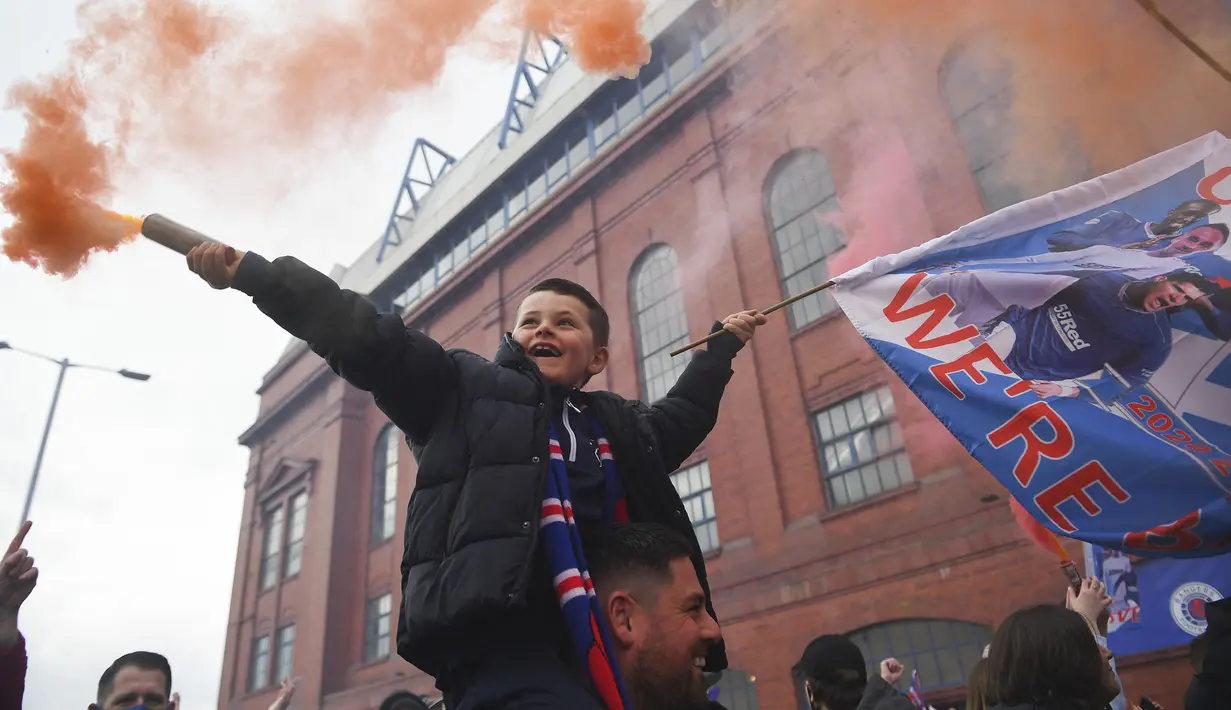 Seorang fans cilik Glasgow Rangers menyalakan granat asap di luar Ibrox Stadium (7/3/2021) untuk merayakan keberhasilan tim kesayangan mereka merebut gelar Liga Utama Skotlandia setelah memimpin 20 poin dari saingan terdekat, Glasgow Celtic. (AFP/Andy Buchanan)