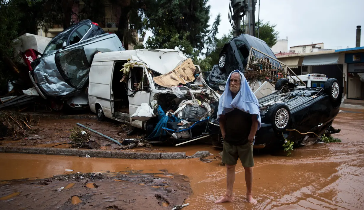 Seorang pria berdiri dekat tumpukan kendaraan yang ringsek akibat diterjang banjir bandang di kota industri Mandra, barat Athena, Rabu (15/11). Menyusul bencana ini, PM Yunani Alexis Tsipras mengumumkan masa berkabung nasional. (AP/Petros Giannakouris)