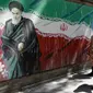 Pejalan kaki melewati lukisan mural yang menggambarkan pendiri Republik Islam Iran Ayatollah Ruhollah Khomeini dan bendera nasional di sepanjang dinding bekas Kedutaan Amerika Serikat (AS) di Ibu Kota Teheran, Iran, Sabtu (22/6/2019). (ATTA KENARE/AFP)