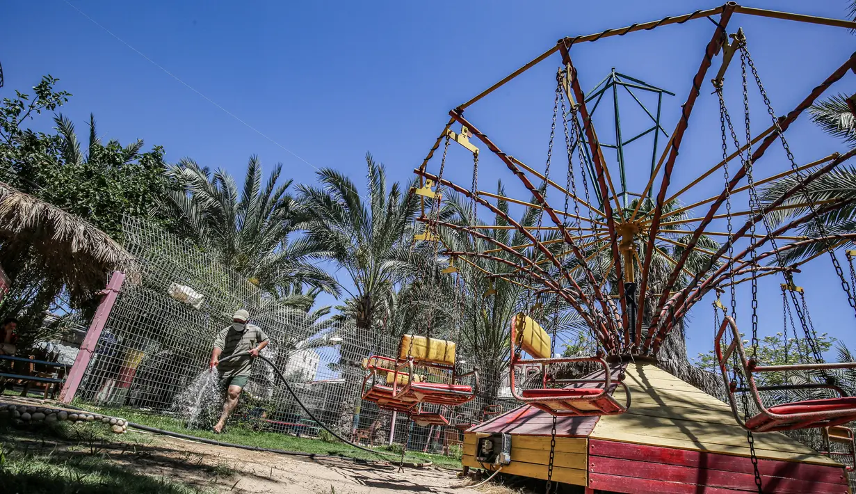 Seorang pekerja mengenakan masker menyiram tanah dan kursi komedi putar di kebun binatang lokal Palestina di Rafah di Jalur Gaza selatan (26/5/2020). Kebun binatang itu ditutup selama liburan Idul Fitri karena pandemi virus coronavirus COVID-19. (AFP/Said Khatib)