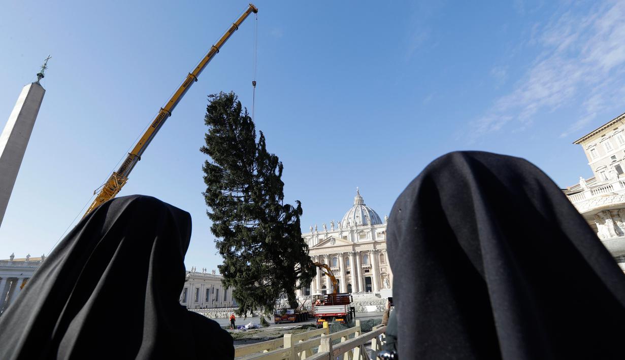 Dua biarawati melihat sebuah derek yang dikerahkan untuk mendirikan pohon Natal raksasa di Alun-alun Santo Petrus, Vatikan, Kamis (23/11). Pohon Natal yang didatangkan dari Polandia ini tingginya mencapai 28 meter. (AP Photo/Andrew Medichini)