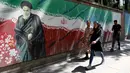 Pejalan kaki melewati lukisan mural yang menggambarkan pendiri Republik Islam Iran Ayatollah Ruhollah Khomeini dan bendera nasional di sepanjang dinding bekas Kedutaan Amerika Serikat (AS) di Ibu Kota Teheran, Iran, Sabtu (22/6/2019). (ATTA KENARE/AFP)