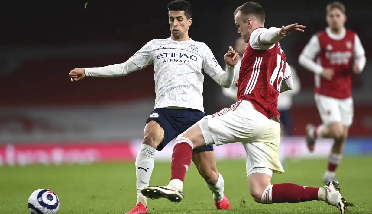 Bek Manchester City, Joao Cancelo berebut bola dengan bek Arsenal, Rob Holding pada lanjutan Liga Inggris di Emirates Stadium, Minggu (21/2/2021). Manchester City kian mengokohkan posisinya di puncak klasemen setelah menang 1-0 di kandang Arsenal. (Shaun Botterill / Pool via AP)