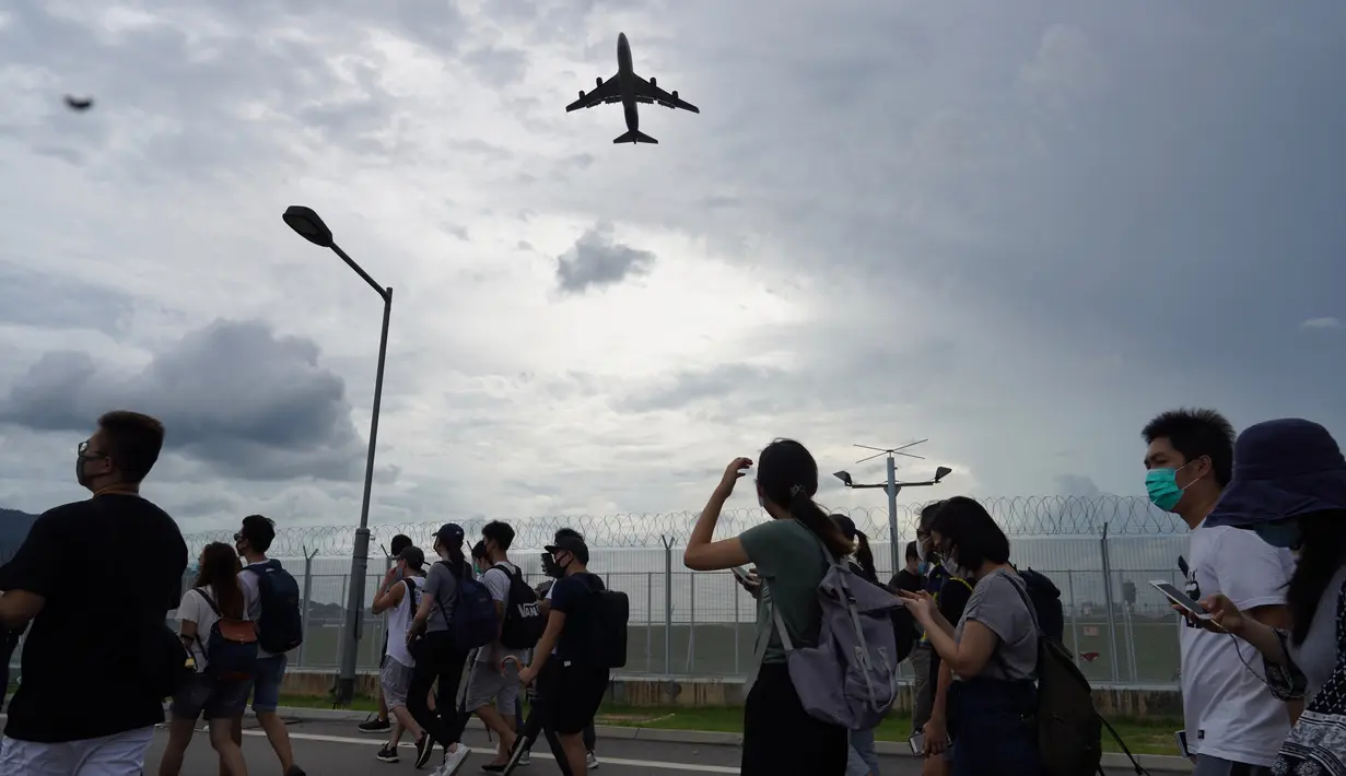 Sebuah pesawat terbang di atas demonstran yang memblokir jalan menuju Bandara Internasional Hong Kong (1/9/2019). Akibat pemblokiran jalan dan layanan kereta api para calon penumpang pesawat terpaksa berjalan kaki menuju bandara. (AP Photo/Vincent Yu)