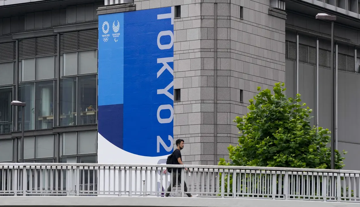 Seseorang dengan masker berjalan di jembatan penyeberangan saat spanduk Tokyo 2020 terlihat di sisi gedung di Tokyo, Rabu (23/6/2021). Olimpiade Tokyo dimulai pada 23 Juli dan penyelenggara mengizinkan penonton lokal menghadiri venue pertandingan dengan kapasitas 50 persen. (AP Photo/Kiichiro Sato)