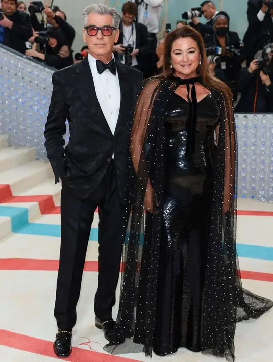 Mantan aktor Bond Pierce Brosnan tak perlu jadi siapa-siapa saat Met Gala. Dalam debutnya, ia mengajak sang istri Keely Shaye yang bersinar dengan gaun cape transparannya.  [Foto: @themetgalaofficial]