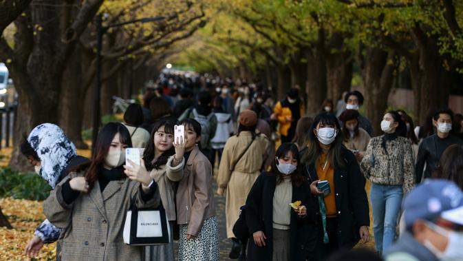 Orang-orang dengan mengenakan masker mengambil foto saat mereka berjalan melalui barisan pohon ginkgo saat pepohonan dan trotoar ditutupi dedaunan kuning cerah di sepanjang trotoar di Tokyo, Jepang pada 28 November 2020. (AP Photo/Kiichiro Sato)