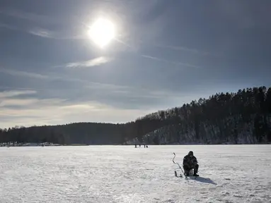 Seorang pria memancing melalui lubang es di danau beku yang tertutup salju dekat Vilnius, Lithuania, Minggu (13/2/2022). Suhu udara 0 derajat Celcius (32 derajat Fahrenheit). (AP Photo/Mindaugas Kulbis)