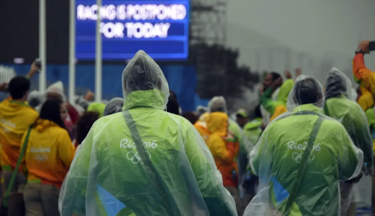 Relawan mengenakan jas hujan sambil mengamati layar billboard yang mengumumkan pembatalan kompetisi cabang dayung Olimpiade 2016 akibat cuaca buruk di stadion Lagoa, Rio de Janeiro, 10 Agustus 2016. (AFP PHOTO/Damien Meyer)