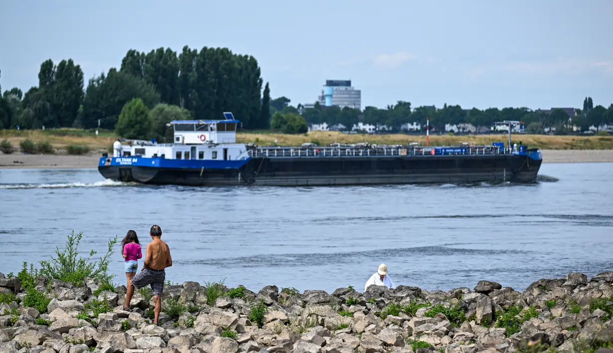 <p>Sebuah kapal pedalaman berlayar di Rhine saat orang-orang berdiri di pantai di tepi sungai yang sebagian mengering di Duesseldorf, Jerman barat (25/7/2022). Keringnya sungah Rhine akibat Eropa mengalami gelombang panas. (AFP/Photo/Ina Fassbender)</p>