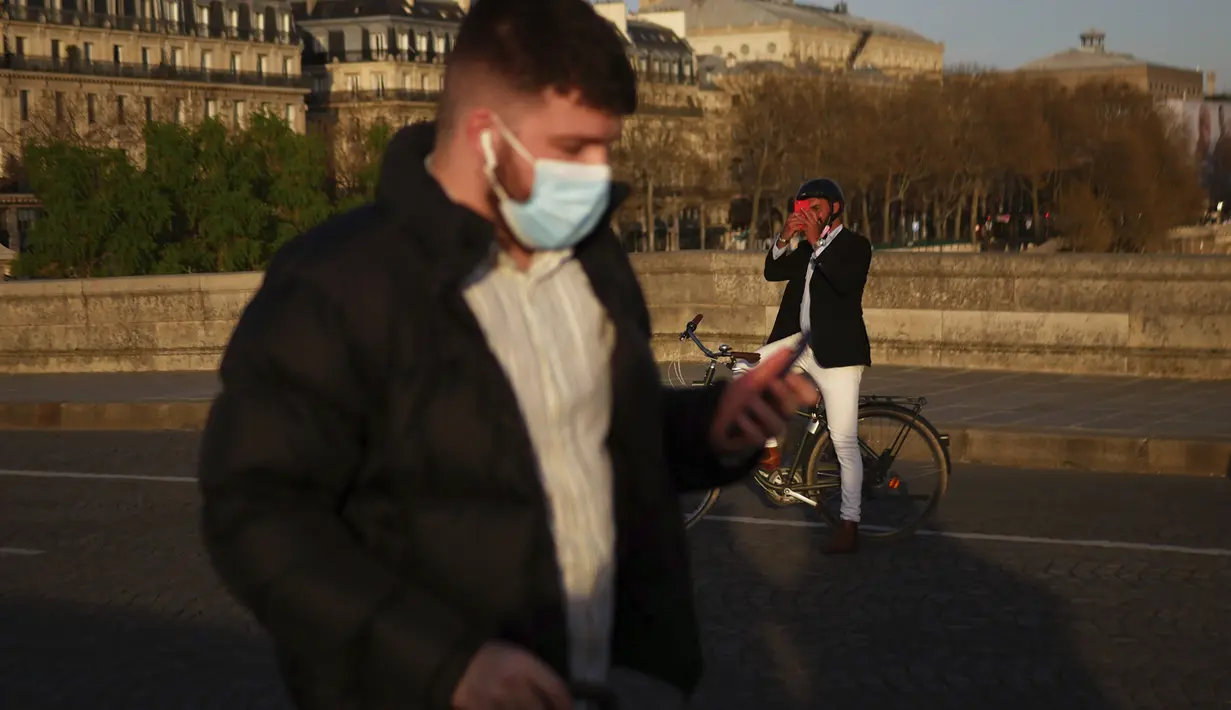 Seorang pria berjalan dengan masker untuk mencegah penyebaran virus corona COVID-19, saat seorang pria mengambil foto matahari terbenam, di Paris (29/3/2021). (AP Photo/Thibault Camus)