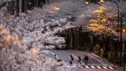 Orang-orang yang mengenakan masker pelindung untuk membantu mengekang penyebaran virus corona berjalan melintasi jalan di bawah bunga sakura di Tokyo, Minggu (28/3/2021). (AP Photo/Kiichiro Sato)