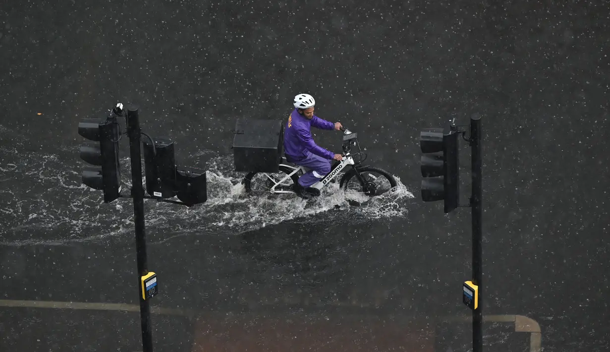 Seorang pengendara sepeda melewati air di jalan yang banjir saat hujan lebat di distrik The Nine Elms London (25/7/2021). Bus dan mobil terjebak banjir ketika jalan-jalan di London banjir pada hari Minggu, ketika badai petir berulang kali melanda ibu kota Inggris. (AFP/Justin Tallis)