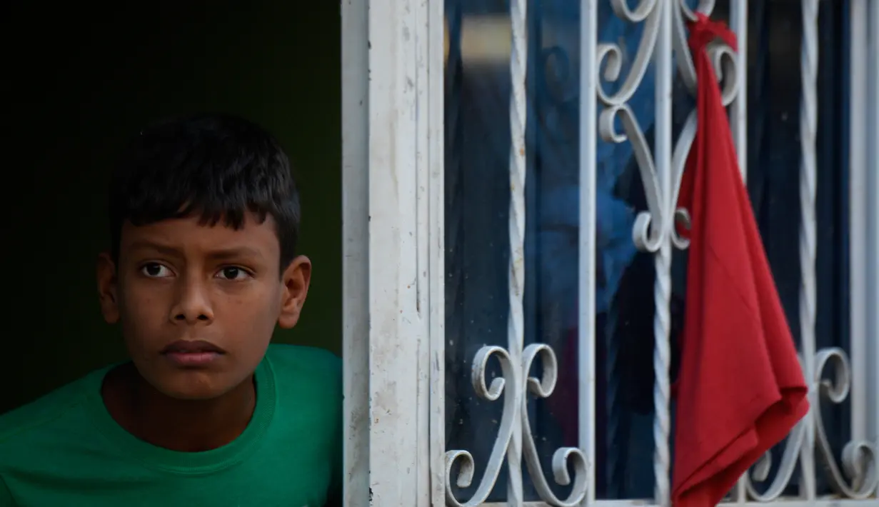 Anak laki-laki memandang ke luar jendela dengan kain merah yang menggantung untuk meminta bantuan makanan selama lockdown di Soacha, dekat Bogota, 4 April 2020. Presiden Kolombia Ivan Duque menetapkan lockdown dari 24 Maret lalu sebagai langkah mencegah penyebaran virus corona. (Raul ARBOLEDA/AFP)