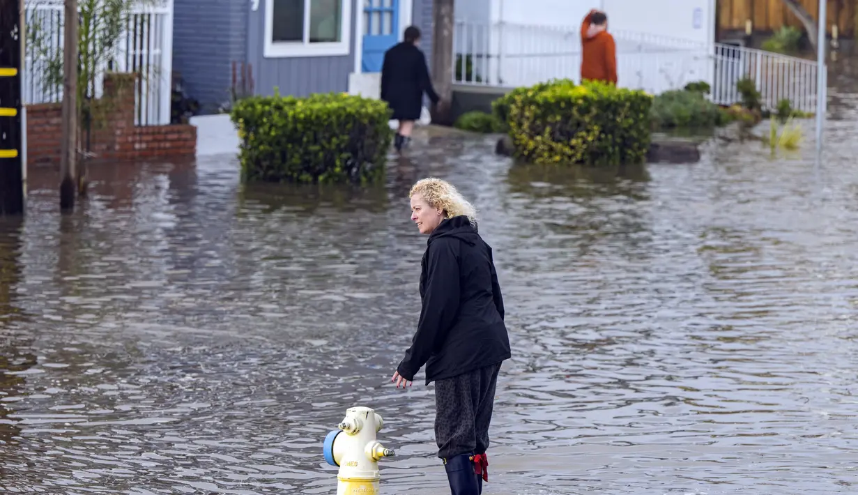 Warga Aptos Beach Flats Dawn Wilkinson berjalan melalui air banjir dari rumahnya di Aptos, California, Kamis (5/1/2023). Angin topan yang merusak, ombak bergelombang dan hujan lebat dari "sungai atmosfer" yang kuat menghantam California pada hari Kamis mengakibatkan kematian setidaknya dua orang, termasuk seorang anak yang rumahnya dihantam pohon tumbang. (AP Photo/Nic Coury)