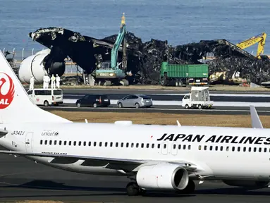 Sebuah pesawat Japan Airlines (JAL) bergerak melewati pekerjaan pemindahan, di bagian belakang, yang sedang berlangsung di lokasi tabrakan pesawat di Bandara Haneda, Tokyo, Jepang, Jumat (5/1/2024). Derek sedang membongkar pesawat Japan Airlines Penerbangan 516 Airbus A350 yang terbakar setelah menabrak pesawat penjaga pantai saat mendarat di bandara. (Kyodo News via AP)
