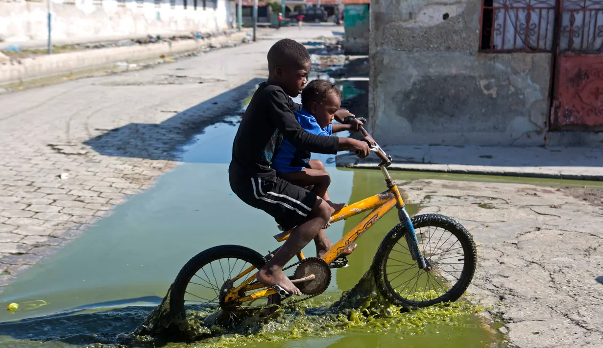 Dua anak melewati genangan air limbah saat naik sepeda melewati daerah Cite Soleil di Port-au-Prince, Haiti (17/3). Cite Soleil awalnya dikembangkan sebagai kota kumuh dan tumbuh hingga sekitar 200.000 hingga 400.000 penduduk. (AP Photo/Dieu Nalio Chery)