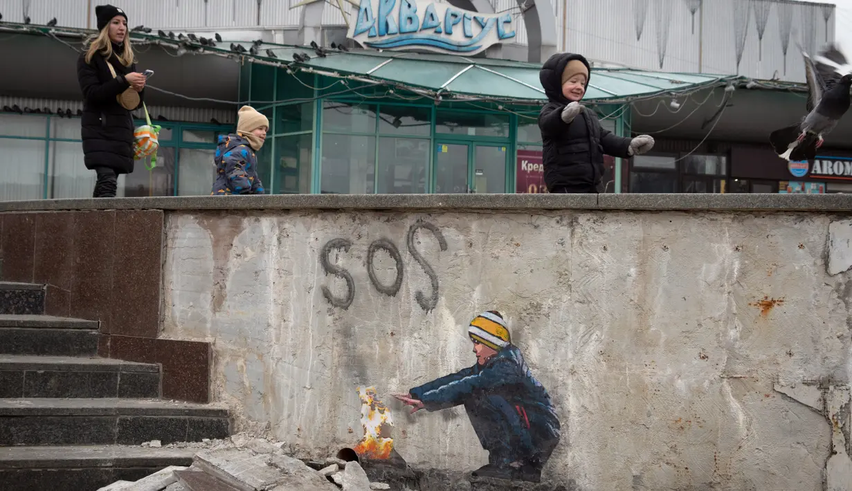 Orang-orang melewati karya seni dari seniman jalanan terkenal TvBoy yang menggambarkan seorang anak laki-laki sedang menghangatkan tangannya dengan api pada dinding yang rusak berat akibat serangan Rusia di Kota Bucha, Ukraina, 30 Januari 2023. Karya seni yang dibuat untuk menghormati para korban perang Rusia-Ukraina tersebut mengandung pesan kelahiran kembali dan harapan serta berharap agar konflik segera berakhir. (AP Photo/Efrem Lukatsky)