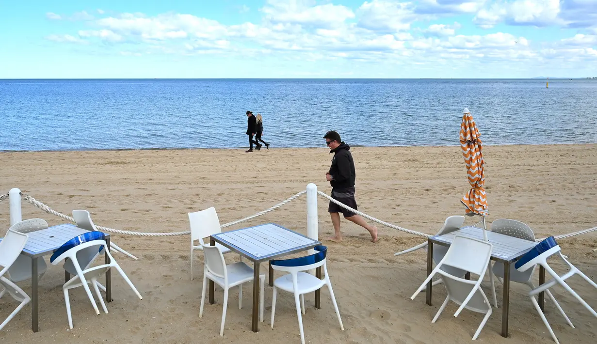 Warga berjalan melewati meja dan kursi kosong di sebuah pantai di Melbourne, Australia, Kamis (19/8/2021). Melbourne melewati 200 hari dalam lockdown keras sejak dimulainya pandemi COVID-19. (William WEST/AFP)