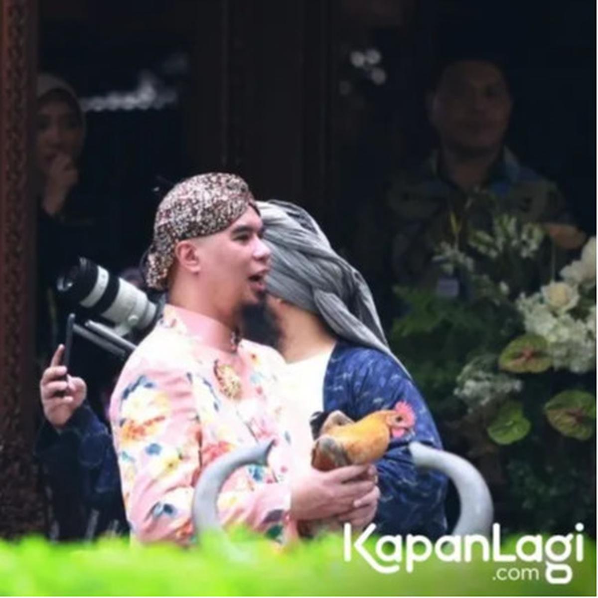 Mengenakan beskap Jawa warna cerah beserta blankon, Ahmad Dhani tampak memegang ayam kampung dengan ekspresi bahagia.