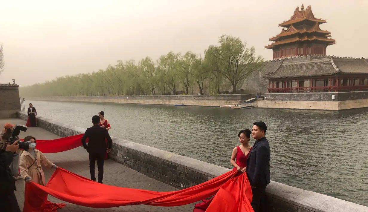 Pasangan pengantin berpose untuk foto di dekat Kota Terlarang saat badai pasir di Beijing (28/3/2021). Badai pasir kembali menyapu Beijing pada Minggu (28/3), mengubah langit menjadi kuning, membatasi jarak pandang dan mengganggu lalu lintas. (AP Photo/Ng Han Guan)