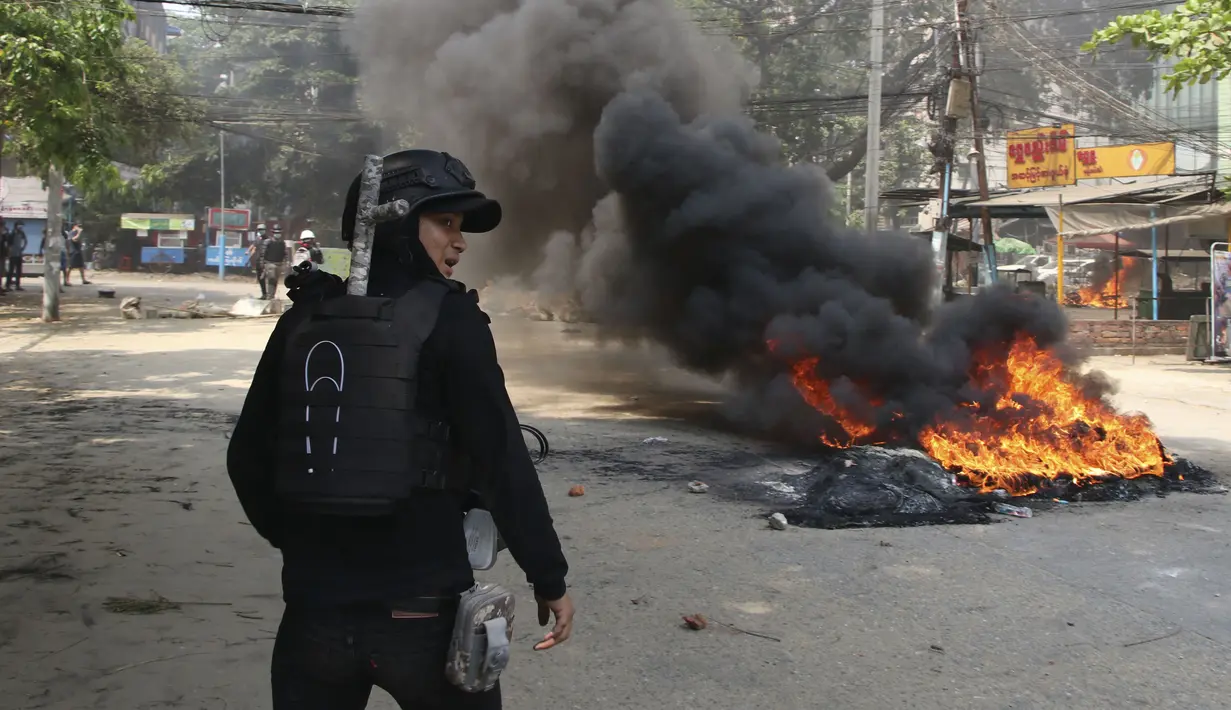 Seorang pengunjuk rasa anti-kudeta berdiri di dekat api selama demonstrasi di kotapraja Dala, Yangon, Myanmar (27/3/2021).  Kepala junta Myanmar menggunakan kesempatan Hari Angkatan Bersenjata untuk mencoba membenarkan penggulingan pemerintahan terpilih Aung San Suu Kyi. (AP Photo)