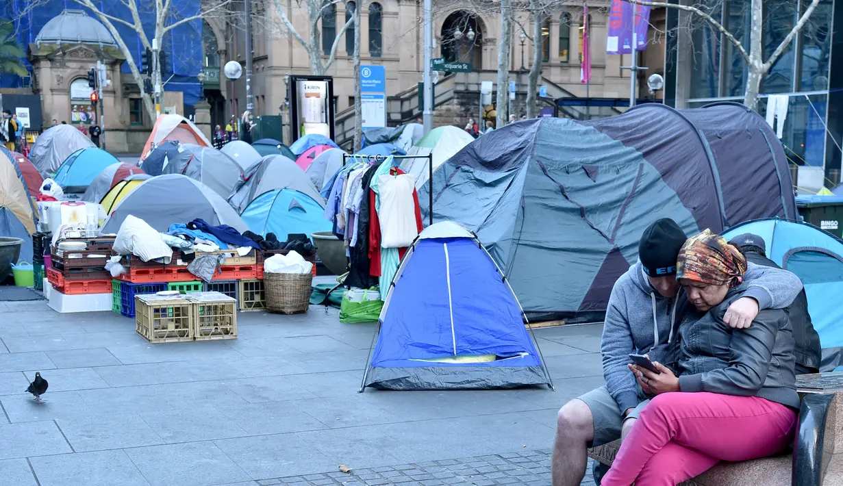 Pasangan tunawisma memainkan ponsel di depan perkampungan tenda para tunawisma di kawasan Martin Place, pusat kota Sydney, Rabu (9/8). Sydney kini tak hanya dipenuhi gedung bertingkat, tetapi juga disesaki tenda-tenda tunawisma. (PETER PARKS / AFP)