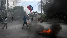 Seorang pria memegang bendera Haiti sementara ban terbakar saat demonstrasi menentang ketidakamanan di Carrefour-Feuilles, Distrik Port-au-Prince, Haiti, 14 Agustus 2023. (Richard PIERRIN/AFP)