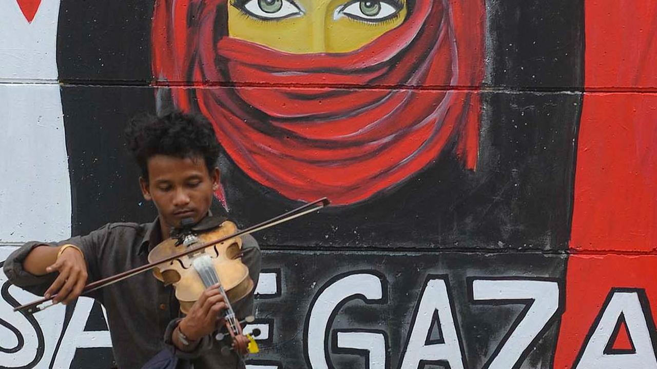 Seorang pengamen memainkan biola di depan mural "Free Palesine' di Jalan Juanda, Kota Depok, Jawa Barat, pada Rabu (8/11/2023).&nbsp;Merdeka.com/Arie Basuki