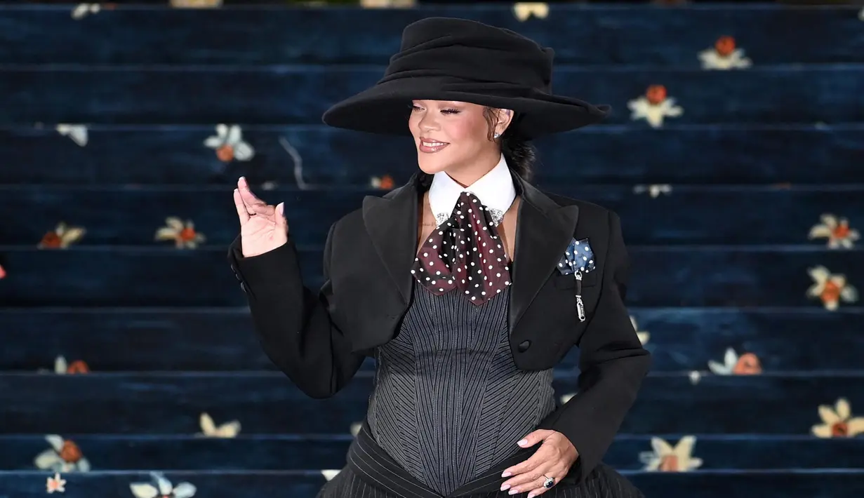 Rihanna tampil sambil memamerkan kehamilannya yang dibalut korset abu-abu dikombinasikan rok hitam selutut dan jaket hitam pendek—semuanya dari Marc Jacobs, yang juga mendesain sepatu hak, dasi, dan topinya (dengan pembuat topi Stephen Jones). (Andrea RENAULT/AFP)
