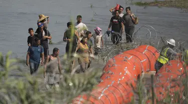 Para migran yang mencoba memasuki AS dari Meksiko mendekati lokasi di mana para pekerja sedang merakit pelampung besar yang akan digunakan sebagai penghalang perbatasan di sepanjang tepi Rio Grande di Eagle Pass, Texas, Selasa (11/7/2023). (AP Photo/Eric Gay)