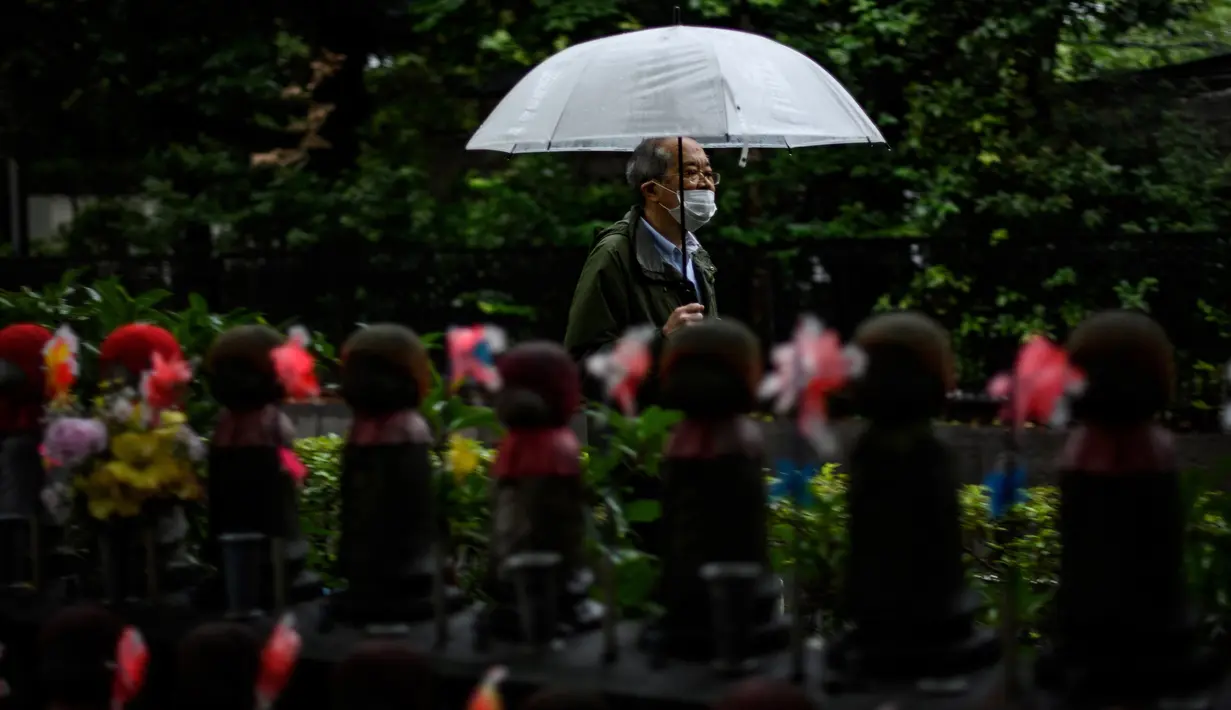 Seorang pria dengan mengenakan payung melintasi patung Jizo (kiri) yang terlihat di Kuil Zojoji, Tokyo pada 22 Juni 2020. Jizo adalah pelindung dari anak-anak yang belum sempat lahir atau meninggal dalam kandungan dan saat masih bayi. (Photo by Philip FONG / AFP)