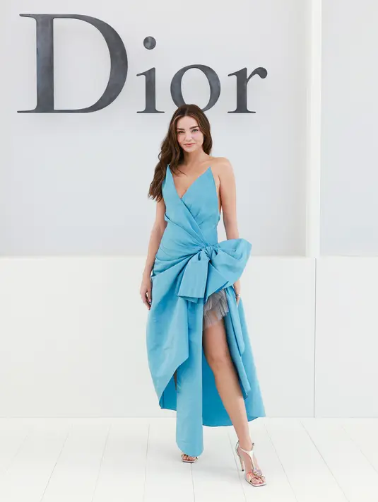 Miranda Kerr tampil dramatis sekaligus playful dengan gaun biru satin asimetris yang dihiasi pita besar di pinggang. Menampilkan rok tulle abu-abu di baliknya, dipadukan heels berhias bunga metalik [dok. Dior]