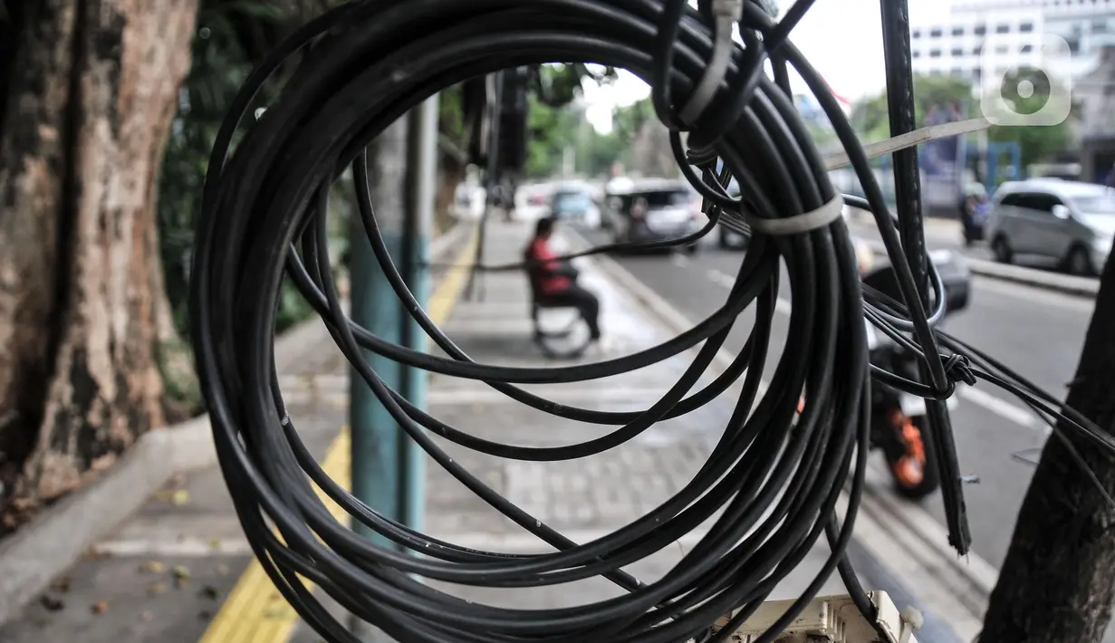 Warga duduk di bawah jaringan kabel utilitas yang menjuntai di Jalan Proklamasi, Pegangsaan, Kecamatan Menteng, Jakarta Pusat, Kamis (6/10/2022). Minimnya pengawasan dalam tahap penyelesaian proyek revitalisasi trotoar oleh pihak terkait menyebabkan jaringan kabel utilitas di kawasan tersebut semrawut  yang dapat membahayakan keselamatan pengguna jalan, terutama saat musim penghujan. (merdeka.com/Iqbal S. Nugroho)