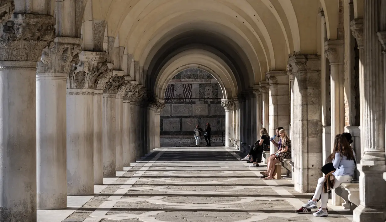 Turis duduk di bawah arcade Istana Doge di Venesia pada 20 Mei 2021. Wisatawan kembali berdatangan menikmati keindahan Venesia, yang sering dijuluki kota paling romantis di dunia, setelah Italia menghapus kewajiban karantina bagi pendatang. (Marco Bertorello/AFP)
