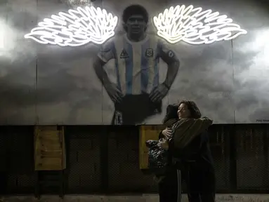 Perempuan berpelukan di dekat mural mendiang legenda sepak bola dunia, Diego Maradona di Buenos Aires, Argentina, Selasa malam (23/11/2021). Peringatan pertama kematian Maradona digelar pada 25 November 2021. (AP Photo/Rodrigo Abd)