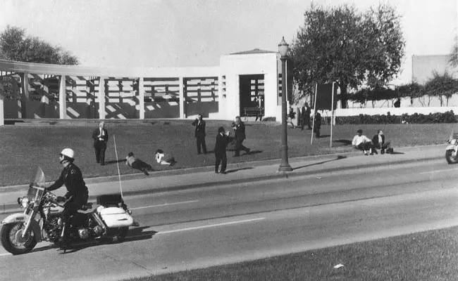 Seorang pria berpayung saat pembunuhan JFK diduga menjadi pelaku. Dalam foto, ia terduduk di paling kanan, dekat rambu. (Sumber Wikimedia Commons/Richard Oscar Bothun untuk ranah publik)