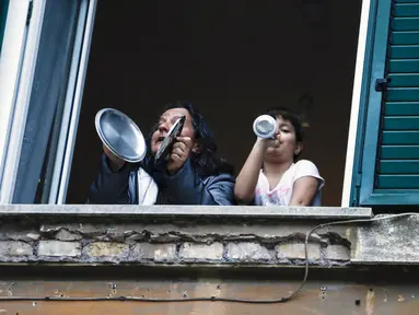 Seorang wanita dan seorang gadis bermain panci dari jendela rumah mereka selama flash mob di lingkungan Garbatella, di Roma, Jumat (13/3/2020). Flash mob ini dilakukan untuk menyatukan orang-orang dan mencoba mengatasi keadaan darurat virus corona Covid-19. (Cecilia Fabiano / LaPresse via AP)