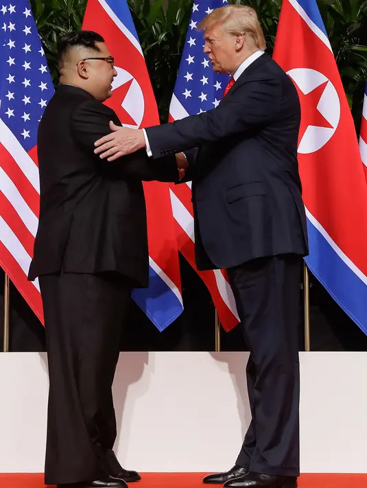 Presiden AS Donald Trump  berjabat tangan dengan Pemimpin Korea Utara, Kim Jong-un dalam pertemuan bersejarah di resor Capella, Pulau Sentosa, Selasa (12/6). Kim dan Trump hadir di depan jurnalis dengan latar belakang bendera Korut dan AS. (AP/Evan Vucci)