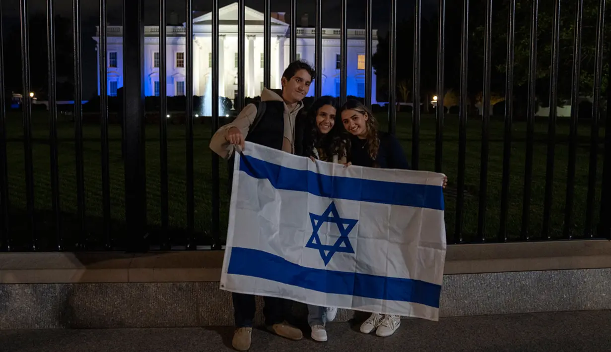 Orang-orang berdiri dengan bendera Israel di luar Gedung Putih yang menyala dengan warna biru dan putih sebagai bentuk solidaritas Amerika Serikat dengan Israel di Washington, Amerika Serikat, Senin (9/10/2023). (AP Photo/Jon Elswick)