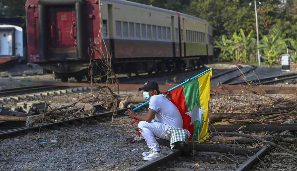 Seorang demonstran membawa bendera nasional Myanmar duduk di rel kereta api yang diblokir dengan kayu, dalam upaya untuk mengganggu layanan kereta selama protes terhadap kudeta militer di Mandalay, Myanmar (17/2/2021). (AP Photo)