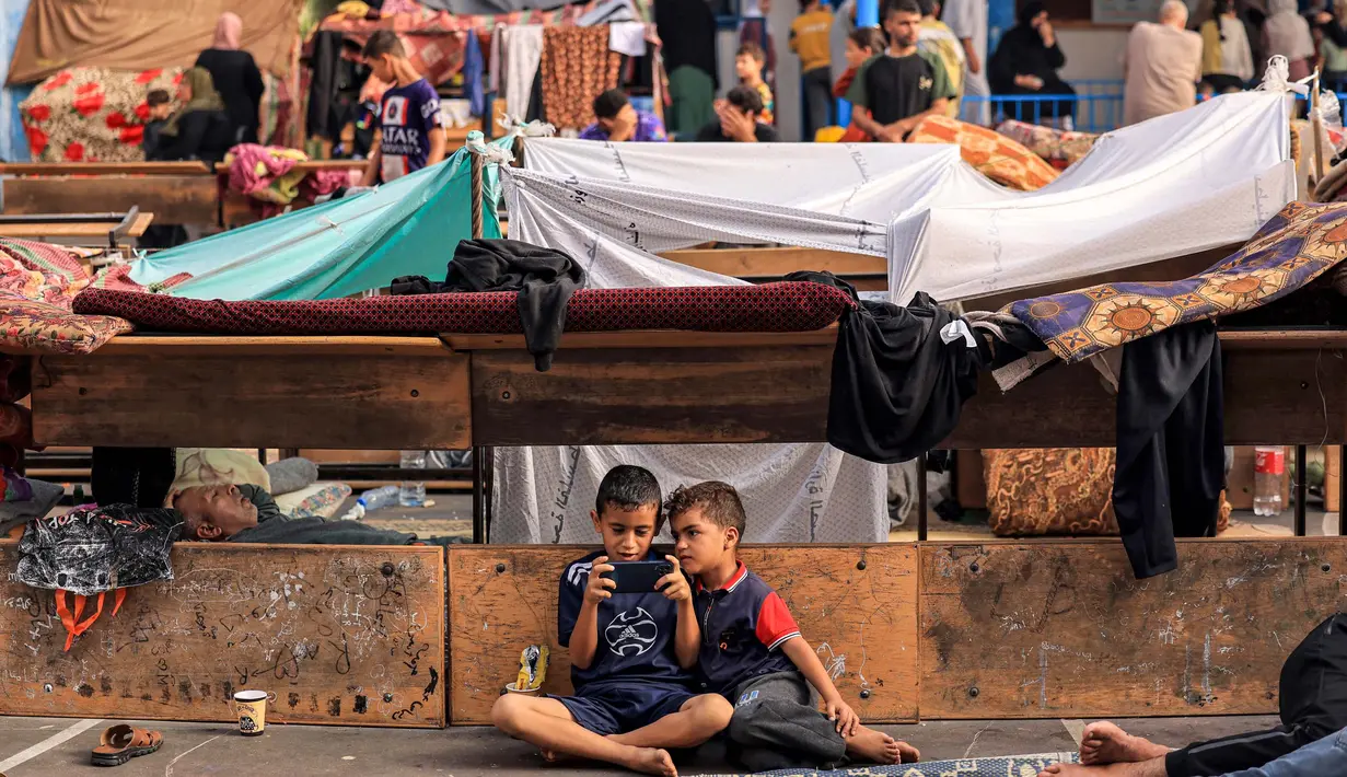 Seorang anak duduk di samping anak lainnya yang sedang bermain ponsel di taman bermain sekolah yang dikelola oleh Badan Bantuan dan Pekerjaan PBB untuk Pengungsi Palestina (UNRWA), 25 Oktober 2023. (MAHMUD HAMS/AFP)