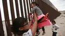 Seorang wanita menemani gadis-gadis kecil bermain jungkat-jungkit yang dipasang di antara pagar pemisah Meksiko dengan Amerika Serikat, Ciudad de Juarez, Meksiko, Minggu (28/7/2019). Jungkat-jungkit tersebut dirancang seorang profesor arsitektur California, Ronald Rael. (AP Photo/Christian Chavez)