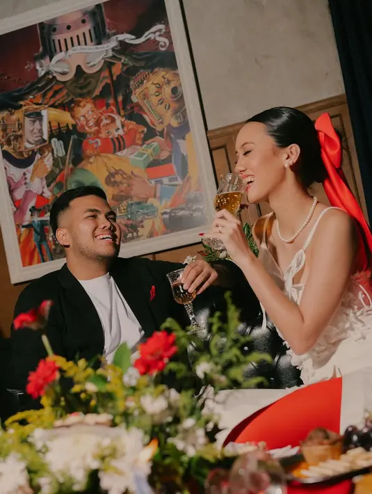 Konsep kedua yang diusung adalah cocktail party. Nabila tampil elegan dengan spaghetti strap dress putih dan aksesori rambut pita merahnya, sedangkan sang suami kenakan setelan jas hitam. [@nocture_co].