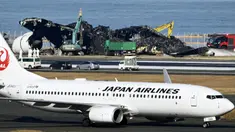 Sebuah pesawat Japan Airlines (JAL) bergerak melewati pekerjaan pemindahan, di bagian belakang, yang sedang berlangsung di lokasi tabrakan pesawat di Bandara Haneda, Tokyo, Jepang, Jumat (5/1/2024). Derek sedang membongkar pesawat Japan Airlines Penerbangan 516 Airbus A350 yang terbakar setelah menabrak pesawat penjaga pantai saat mendarat di bandara. (Kyodo News via AP)
